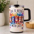 A handled insulated travel tumbler with a clear lid and metal straw, featuring a colorful watercolor-style illustration of six cats and the text 'LIFE IS BETTER WITH CATS.' The tumbler is shown standing on a light surface with a blurred plant and candle in the background.