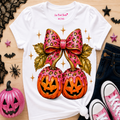 A white graphic t-shirt featuring a pink leopard-print bow and two pink-dripped jack-o'-lanterns, styled in a flat lay with jeans, pink sneakers, and Halloween decorations on a wooden surface.