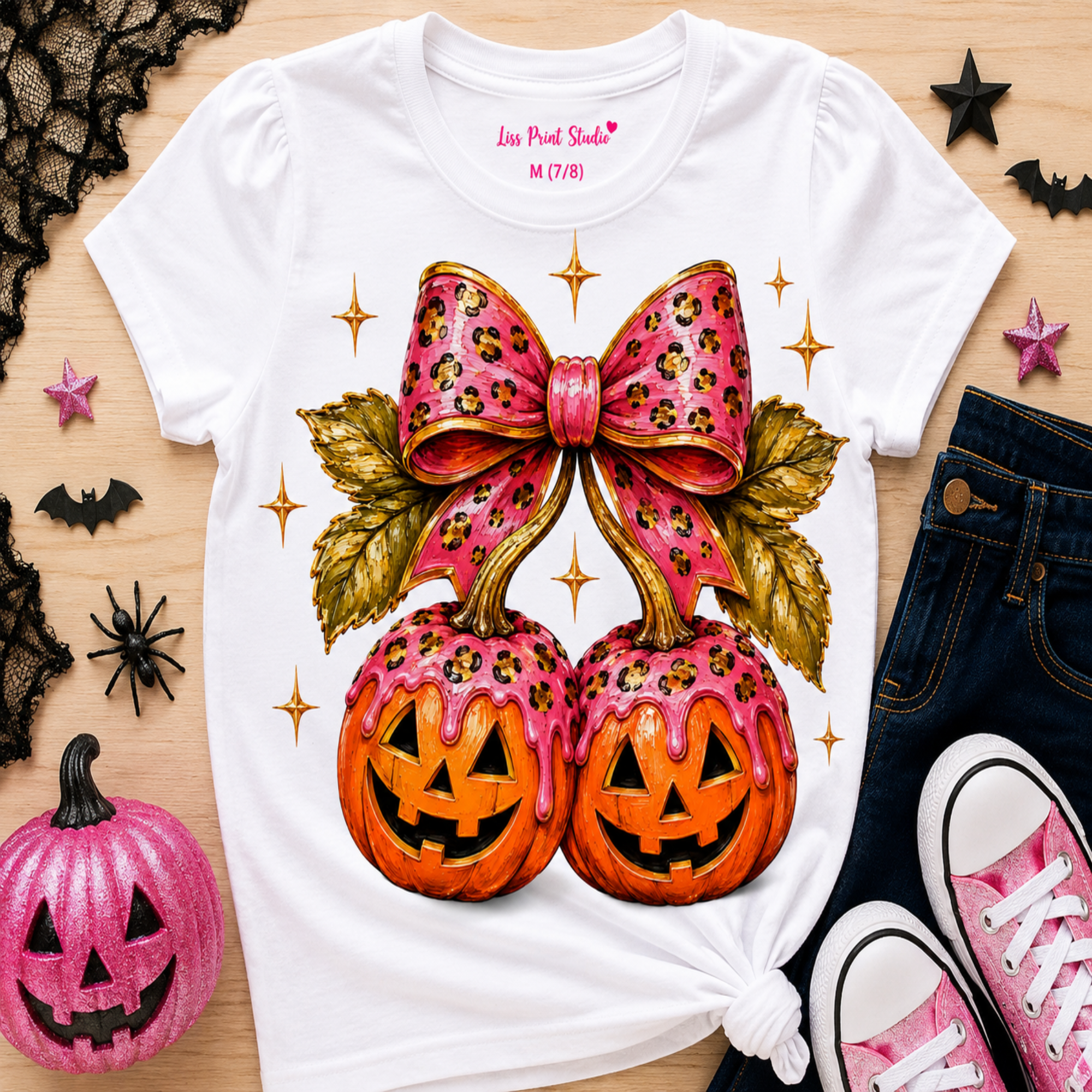 A white graphic t-shirt featuring a pink leopard-print bow and two pink-dripped jack-o'-lanterns, styled in a flat lay with jeans, pink sneakers, and Halloween decorations on a wooden surface.