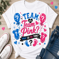 A white graphic T-shirt laid flat on a light wood surface, surrounded by white sneakers, blue jeans, and small pink and blue heart decorations. The shirt graphic reads “TEAM or Team Pink? WHAT DO YOU THINK?” with blue and pink balloons, baby onesies, stars, and footprints.