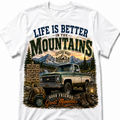 A white short-sleeve crewneck t-shirt featuring a large graphic print on the front. The graphic depicts a vintage green pickup truck in a mountain landscape with pine trees, a campsite signpost, and text reading 'LIFE IS BETTER IN THE MOUNTAINS' and 'GOOD FRIENDS Great Memories'.