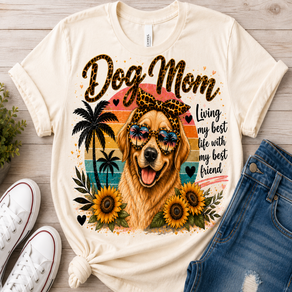 A cream-colored graphic t-shirt featuring a golden retriever dog wearing sunglasses, surrounded by palm trees, sunflowers, and the text 'Dog Mom' and 'Living my best life with my best friend'. The shirt is styled in a flat-lay arrangement on a light wood background alongside white sneakers and blue jeans.