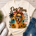 A cream-colored graphic t-shirt featuring a golden retriever dog wearing sunglasses, surrounded by palm trees, sunflowers, and the text 'Dog Mom' and 'Living my best life with my best friend'. The shirt is styled in a flat-lay arrangement on a light wood background alongside white sneakers and blue jeans.