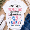 A white graphic t-shirt laid flat on a wooden surface, styled with white sneakers, blue jeans, and a knit sweater. The shirt features a colorful print that reads 'WE'RE EXPECTING OUR GREATEST Adventure BABY EST.