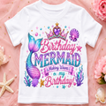 A white short-sleeve kids' t-shirt with a colorful mermaid-themed birthday graphic, laid flat on a pink fabric background decorated with flowers and starfish.