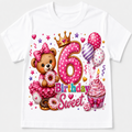A white short-sleeve kids' t-shirt featuring a colorful '6th Birthday' graphic print with a teddy bear, donuts, balloons, a cupcake, and a gold crown.
