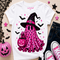 A white graphic T-shirt laid flat on a light wood surface, featuring a pink witch ghost design with a black hat, pink pumpkin, bats, and stars. The shirt is styled with a pink jack-o'-lantern, dark jeans, pink sneakers, black lace, and star decorations.