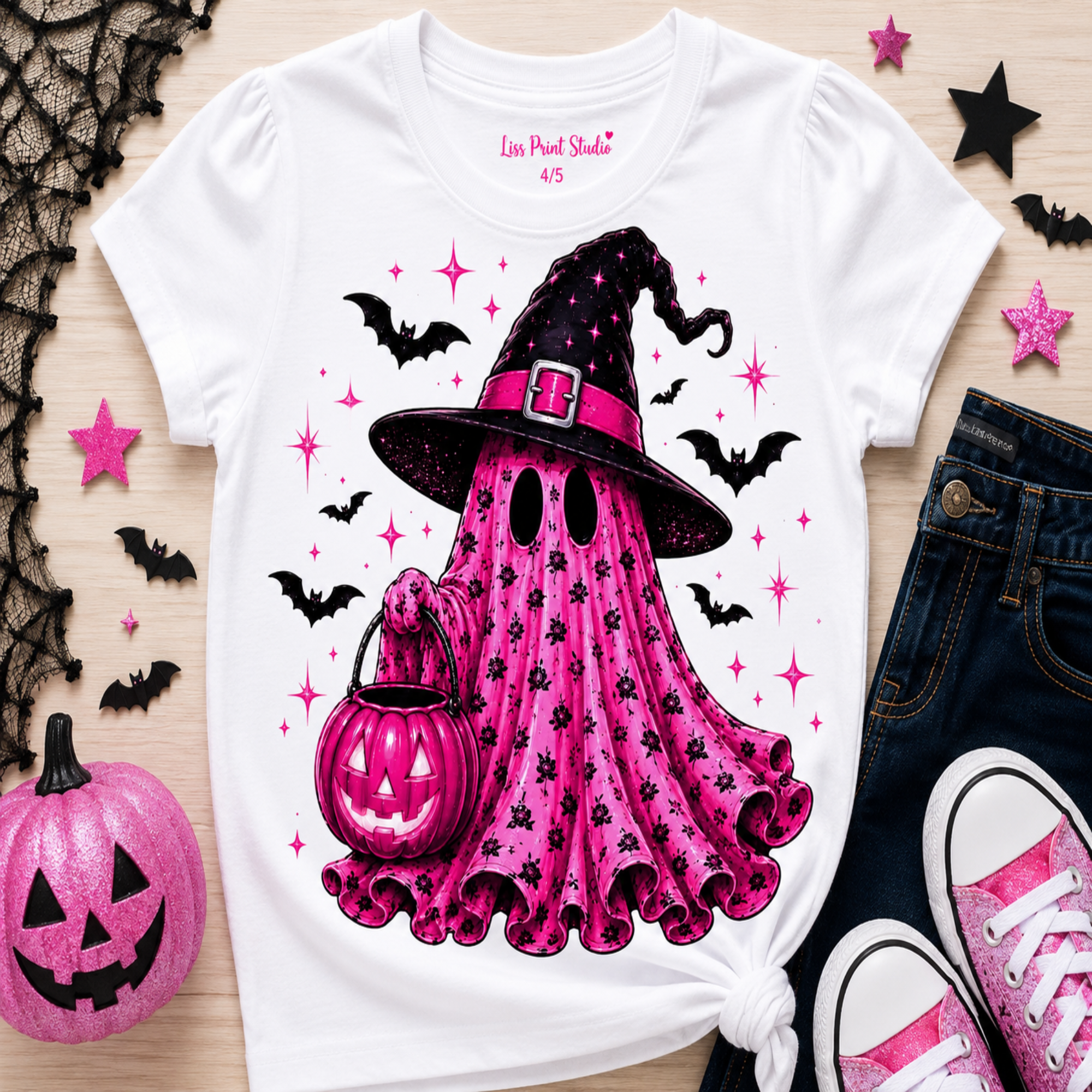 A white graphic T-shirt laid flat on a light wood surface, featuring a pink witch ghost design with a black hat, pink pumpkin, bats, and stars. The shirt is styled with a pink jack-o'-lantern, dark jeans, pink sneakers, black lace, and star decorations.