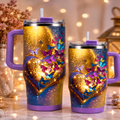 Two handled insulated tumblers with lids and straws, shown on a tabletop in a warm indoor setting. The tumblers have a printed design featuring a large glowing gold heart, smaller gold hearts, colorful butterflies, and a blue-to-purple gradient background with sparkles.