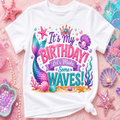 A white kids' t-shirt with a colorful mermaid-themed birthday graphic that reads 'It's My BIRTHDAY! Let's Make Some WAVES!'. The shirt is styled in a flat-lay on a pink textured background with various mermaid-themed props including starfish, shells, pearls, and sequins.