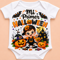 White short-sleeve baby bodysuit with a colorful Halloween graphic print featuring the Spanish text 'Mi Primer HALLOWEEN', a cartoon child in a witch outfit, pumpkins, bats, a ghost, and a haunted house, shown on a peach background.