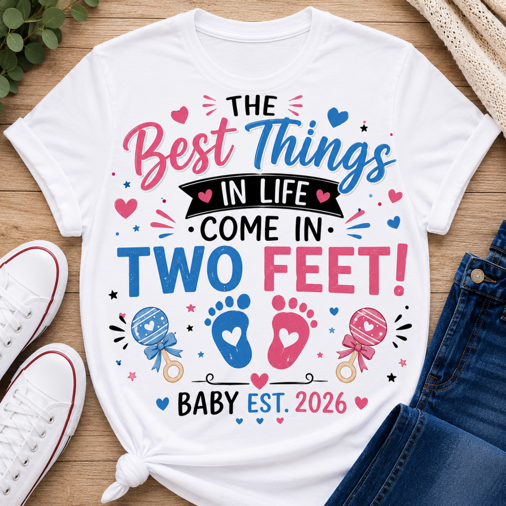 A white graphic t-shirt laid flat on a wooden surface, styled with white sneakers, blue jeans, and a small plant. The t-shirt features a colorful graphic that reads 'THE Best Things IN LIFE COME IN TWO FEET! BABY EST.