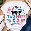 A white graphic t-shirt laid flat on a wooden surface, styled with white sneakers, blue jeans, and a small plant. The t-shirt features a colorful graphic that reads 'THE Best Things IN LIFE COME IN TWO FEET! BABY EST.