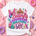 A white short-sleeve T-shirt with a colorful 'Butterfly Birthday Girl' graphic, styled in a flat lay on a pink background with flowers and beads.