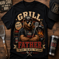 A black graphic T-shirt laid flat on a wooden surface, featuring a bear wearing a BBQ cap and apron with the words 'GRILL' and 'FATHER' in large red and beige lettering. Surrounding props include a metal spatula, a matching cap, a tumbler, and a sign reading 'MAN CAVE EAT.