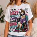 A woman wearing a white graphic t-shirt with a colorful cartoon cat sitting in a teal chair, wearing sunglasses and a leopard print hat. The text on the shirt reads 'I'M NOT LAZY I'M JUST IN Energy SAVING MODE DO NOT DISTURB MY PEACE' and other motivational phrases.