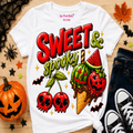 A white graphic T-shirt laid flat on a wood surface with Halloween-themed props including a jack-o'-lantern, jeans, sneakers, spiderwebs, and autumn leaves. The shirt features large red and green text reading 'Sweet & Spooky' above illustrated cherries, a watermelon ice cream cone, bats, and stars.