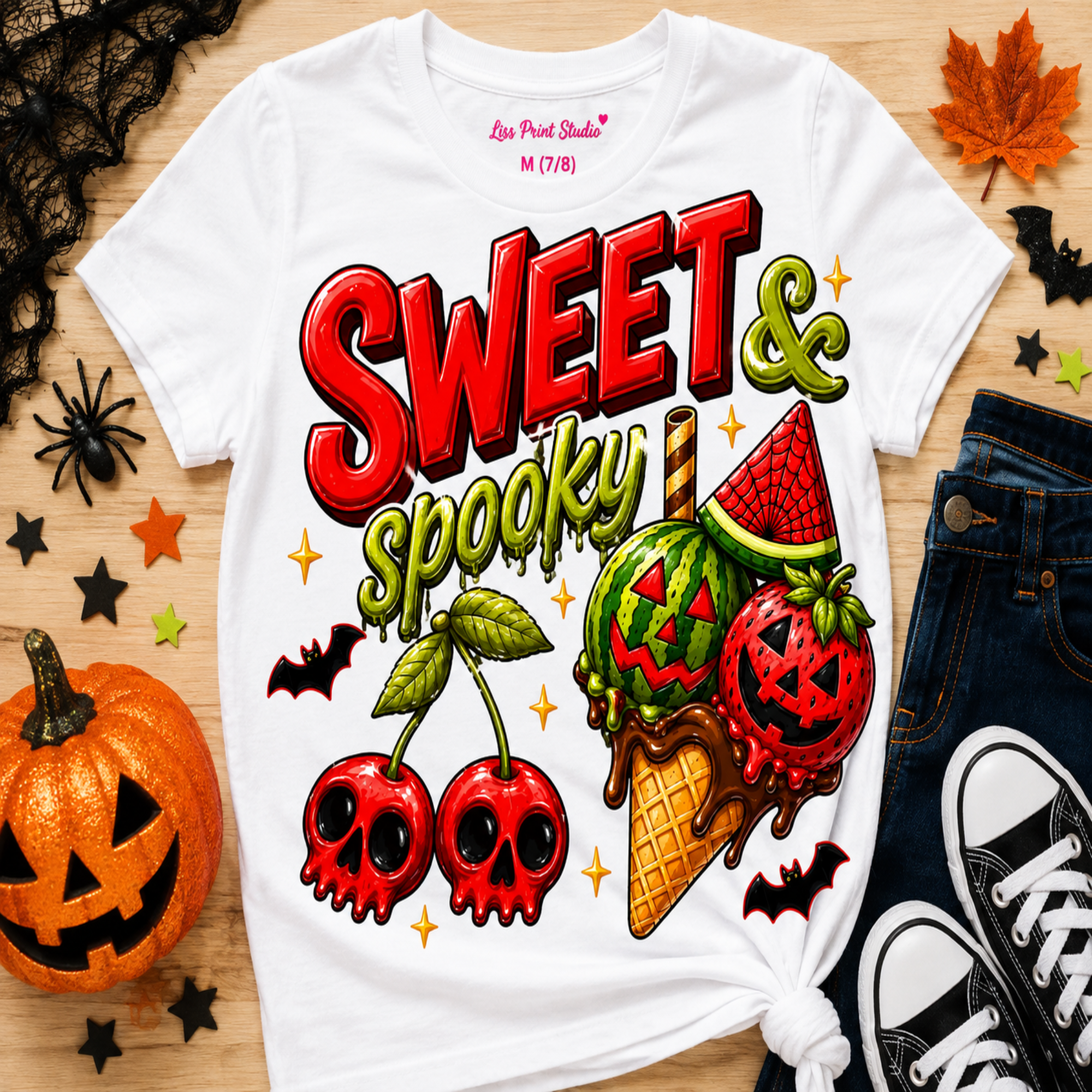 A white graphic T-shirt laid flat on a wood surface with Halloween-themed props including a jack-o'-lantern, jeans, sneakers, spiderwebs, and autumn leaves. The shirt features large red and green text reading 'Sweet & Spooky' above illustrated cherries, a watermelon ice cream cone, bats, and stars.