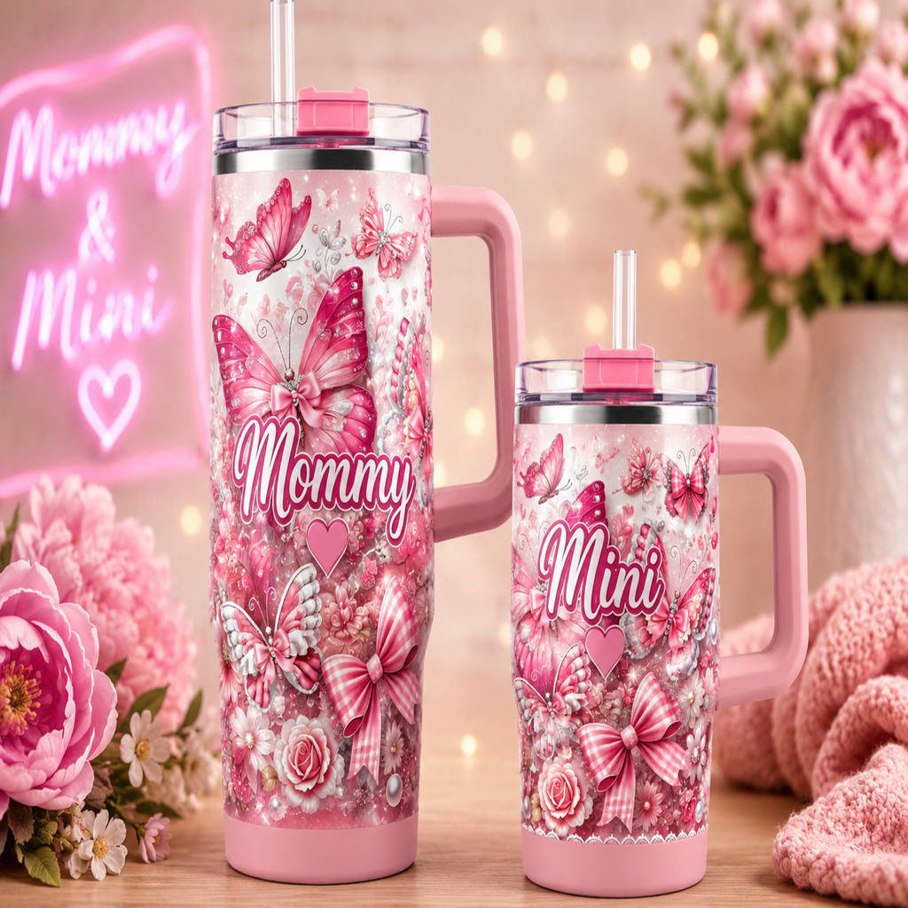 Two pink handled tumblers with clear lids and straws, decorated with butterflies, bows, roses, and the words 'Mommy' and 'Mini,' shown on a tabletop with flowers, a pink neon sign, and warm string lights in the background.