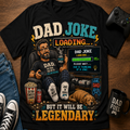 A black graphic t-shirt laid flat on a wooden surface, featuring a humorous 'Dad Joke Loading' design with a cartoon dad, a dog, a popcorn bucket, and gaming elements. The shirt is styled with jeans, sneakers, a game controller, and a mug.