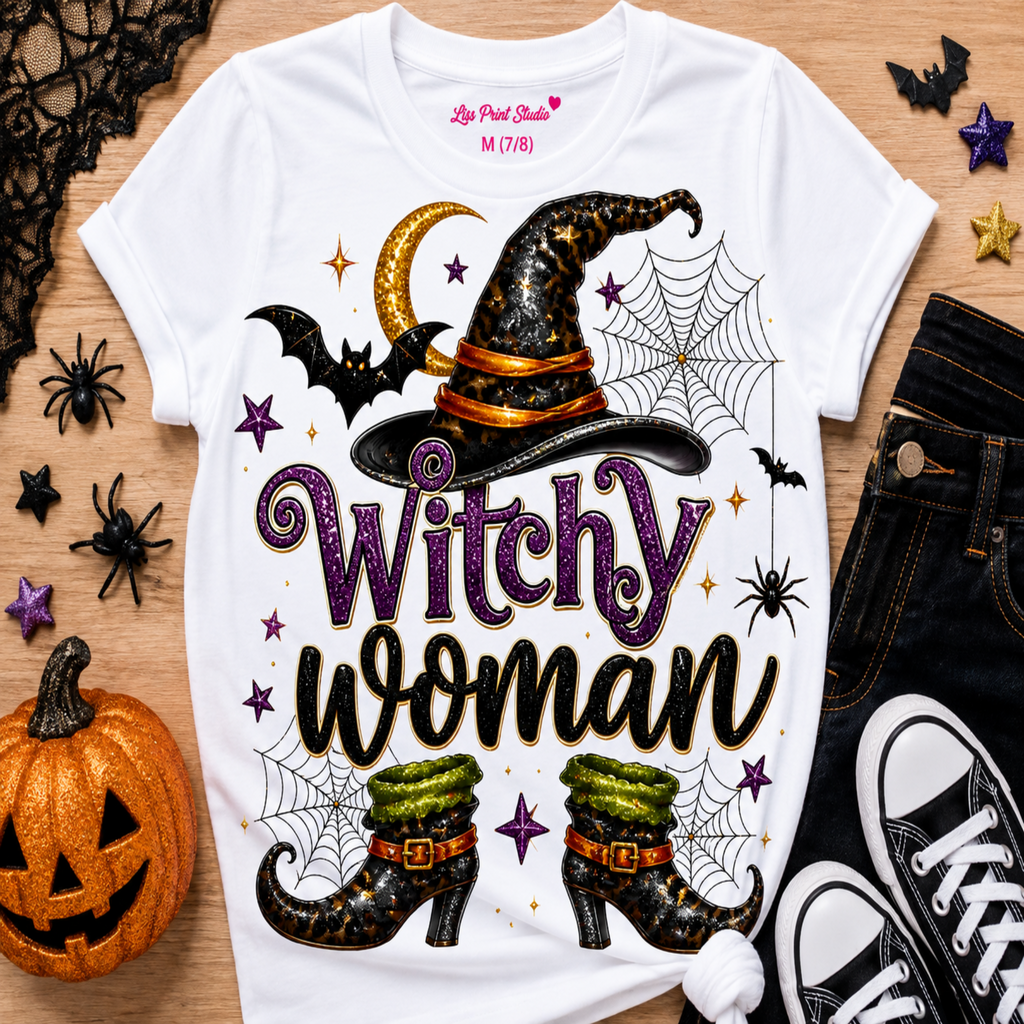 A white graphic T-shirt laid flat on a wood surface with Halloween props around it. The shirt features a witch hat, spiderwebs, bats, stars, and the words 'Witchy woman' in purple and black lettering.