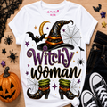 A white graphic T-shirt laid flat on a wood surface with Halloween props around it. The shirt features a witch hat, spiderwebs, bats, stars, and the words 'Witchy woman' in purple and black lettering.