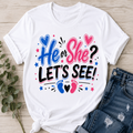 White graphic T-shirt laid flat on a light wood surface with a knot at the bottom, styled with blue jeans and a small green sprig. The shirt reads “He or She? LET’S SEE!” in blue and pink lettering with hearts, stars, and baby footprints.