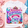 A white short-sleeve t-shirt with a colorful mermaid-themed 'Mermaid Birthday Too Magical to be ONE' graphic, styled in a flat-lay on a pink background with seashells, starfish, and pearls.