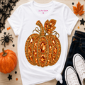 A white graphic t-shirt featuring a large, ornate glittery orange pumpkin design on the front. The t-shirt is styled in a flat-lay arrangement on a light wood background with autumn props including a real pumpkin, a black spider decoration, a black lace piece, a maple leaf, and dark blue jeans with white sneakers.