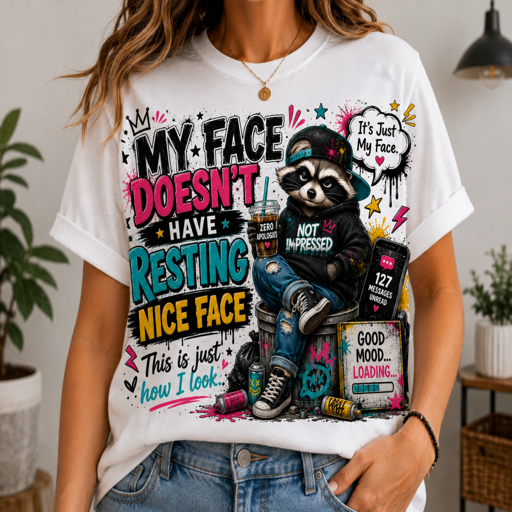 A woman wearing a white graphic t-shirt with a humorous raccoon design. The graphic features a cartoon raccoon wearing a cap and hoodie sitting on a pile of junk, with text that reads 'MY FACE DOESN'T HAVE RESTING NICE FACE' and 'This is just how I look.