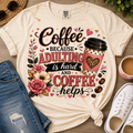 A cream-colored graphic t-shirt styled in a flat-lay arrangement with blue jeans, white sneakers, sunglasses, and a cup of coffee on a wooden surface. The t-shirt features a graphic that reads 'Coffee BECAUSE ADULTING is hard AND COFFEE helps' with a coffee cup illustration and floral details.