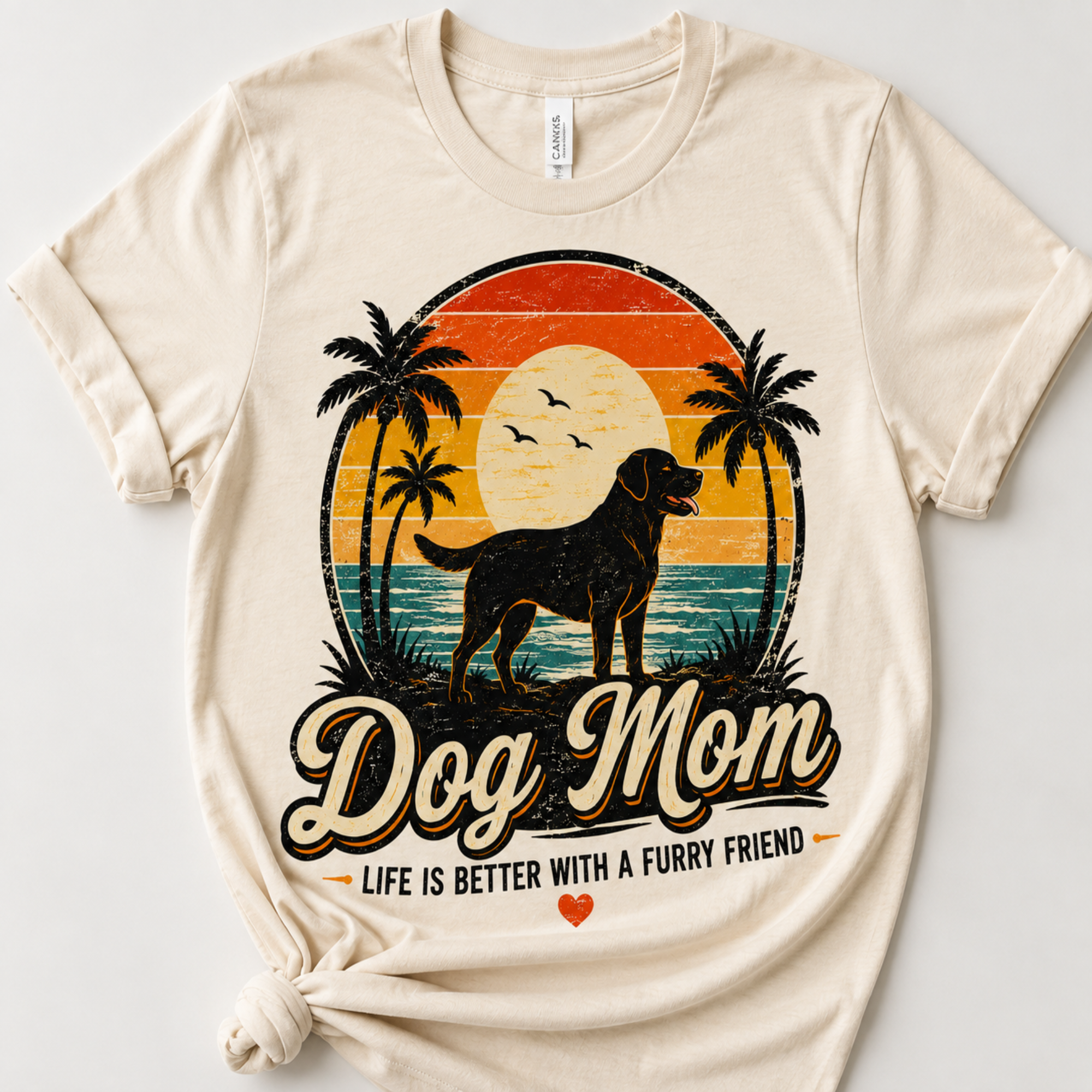 A beige short-sleeve t-shirt with a distressed retro graphic of a dog standing in front of a sunset and palm trees, with the text 'Dog Mom' and 'LIFE IS BETTER WITH A FURRY FRIEND'.