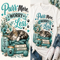 A white graphic T-shirt shown in a styled flat-lay with jeans, white sneakers, flowers, and a mug. The shirt features a large printed design with the phrase 'Purr More Worry Less,' a sleeping tabby kitten on a teal knit blanket, flowers, a mason jar, books, a cup with a paw print, and small decorative elements.