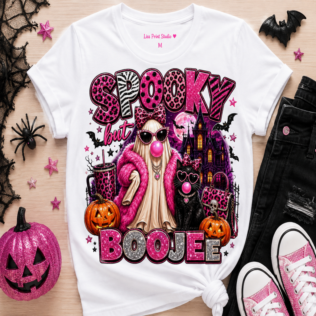 A white graphic t-shirt featuring a 'Spooky But Boojee' Halloween design with a pink nose, cat, pumpkins, and leopard print lettering, styled in a flat lay with black jeans, pink sneakers, and a glittery jack-o'-lantern on a light wood background.