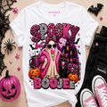 A white graphic t-shirt featuring a 'Spooky But Boojee' Halloween design with a pink nose, cat, pumpkins, and leopard print lettering, styled in a flat lay with black jeans, pink sneakers, and a glittery jack-o'-lantern on a light wood background.