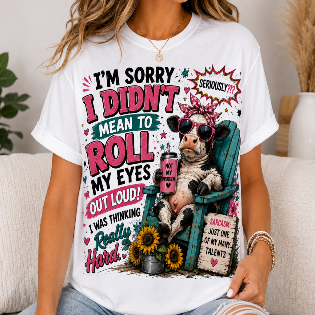 A woman wearing a white graphic t-shirt with a humorous cartoon design of a cow in sunglasses sitting in a teal chair, surrounded by text that reads 'I'M SORRY I DIDN'T MEAN TO ROLL MY EYES OUT LOUD!'.