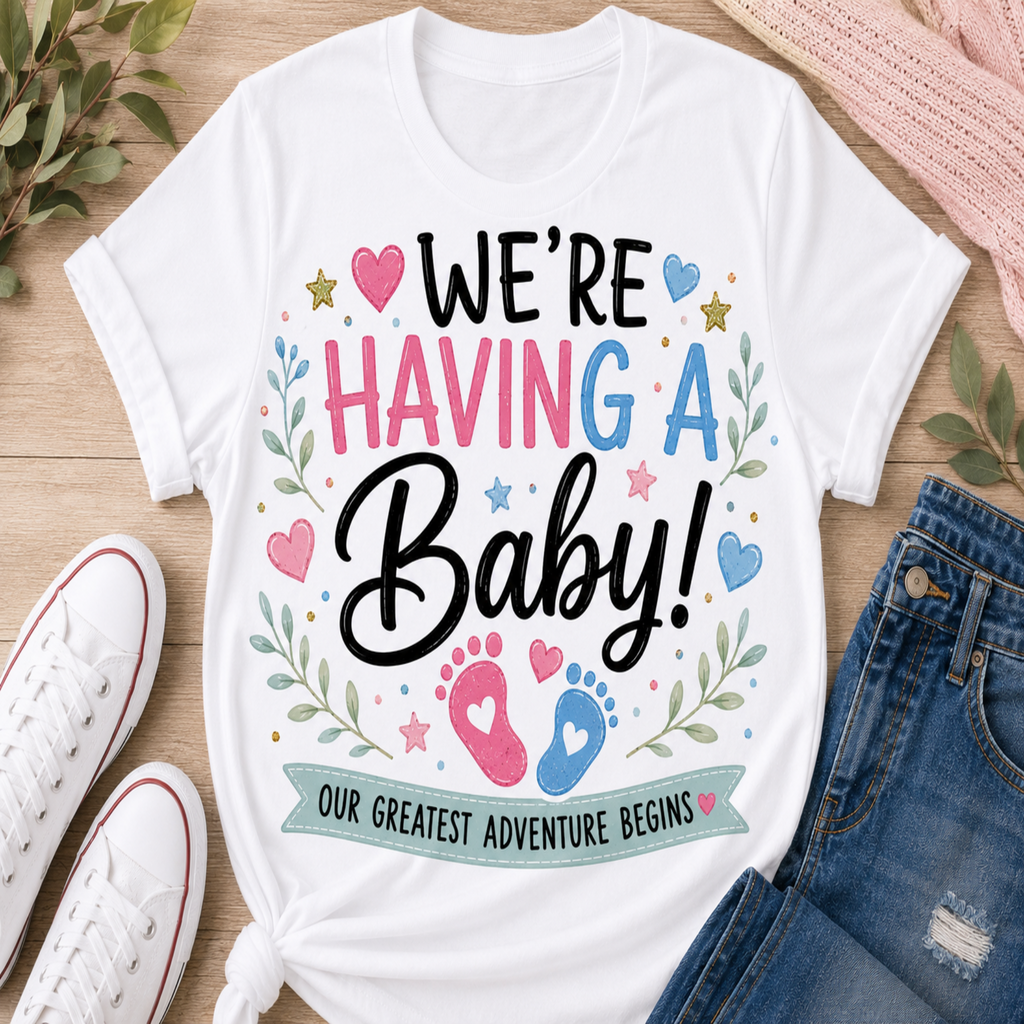 A white graphic t-shirt laid flat on a light wood surface, surrounded by white sneakers, blue jeans, and green leaves. The t-shirt features a colorful design that reads 'WE'RE HAVING A Baby! OUR GREATEST ADVENTURE BEGINS' with pink and blue baby footprints and hearts.