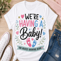 A white graphic t-shirt laid flat on a light wood surface, surrounded by white sneakers, blue jeans, and green leaves. The t-shirt features a colorful design that reads 'WE'RE HAVING A Baby! OUR GREATEST ADVENTURE BEGINS' with pink and blue baby footprints and hearts.