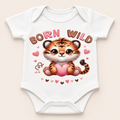 A white short-sleeve baby bodysuit with a cartoon tiger graphic holding a pink heart, surrounded by the words 'BORN WILD' and small pink hearts, shown on a light beige background.