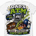 A white short-sleeve t-shirt featuring a colorful cartoon graphic of an ATM machine with a mustache, surrounded by money, a crying wallet, a blue backpack, and bold text reading 'DAD'S ATM' and 'KIDS APPROVED SINCE DAY ONE'.