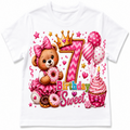 A white short-sleeve kids' t-shirt featuring a colorful, detailed graphic design for a 7th birthday. The design includes a brown teddy bear in a pink dress holding a donut, a large pink and gold number 7 with a crown, pink balloons, a pink cupcake with a candle, and the words 'Birthday Sweet'.