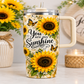 A handleless tumbler with a lid and straw, decorated with yellow sunflowers, green leaves, and butterflies, and the phrase 'You are my Sunshine' in black script. The tumbler is shown on a tabletop with sunflowers and a lit candle in the background.