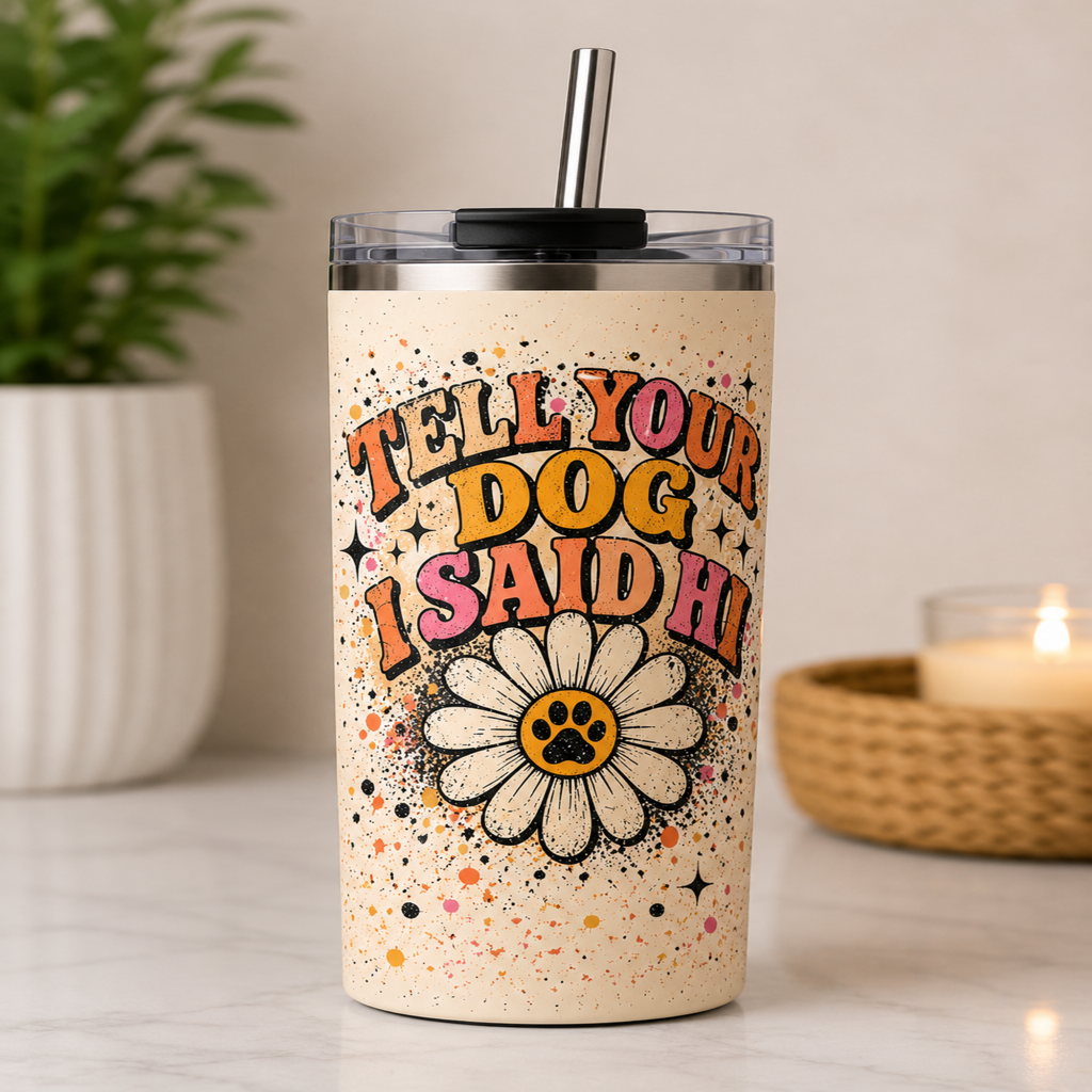 A stainless steel insulated tumbler with a clear lid and metal straw, featuring the printed phrase “TELL YOUR DOG I SAID HI” in colorful retro lettering above a daisy with a paw print center. The tumbler is photographed on a marble surface with a potted plant and a lit candle in the background.