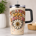A stainless steel handled tumbler with a clear lid and straw, featuring a beige speckled background and the text 'TELL YOUR DOG I SAID HI' above a daisy flower with a paw print center. The tumbler is shown on a marble countertop with a blurred plant and candle in the background.