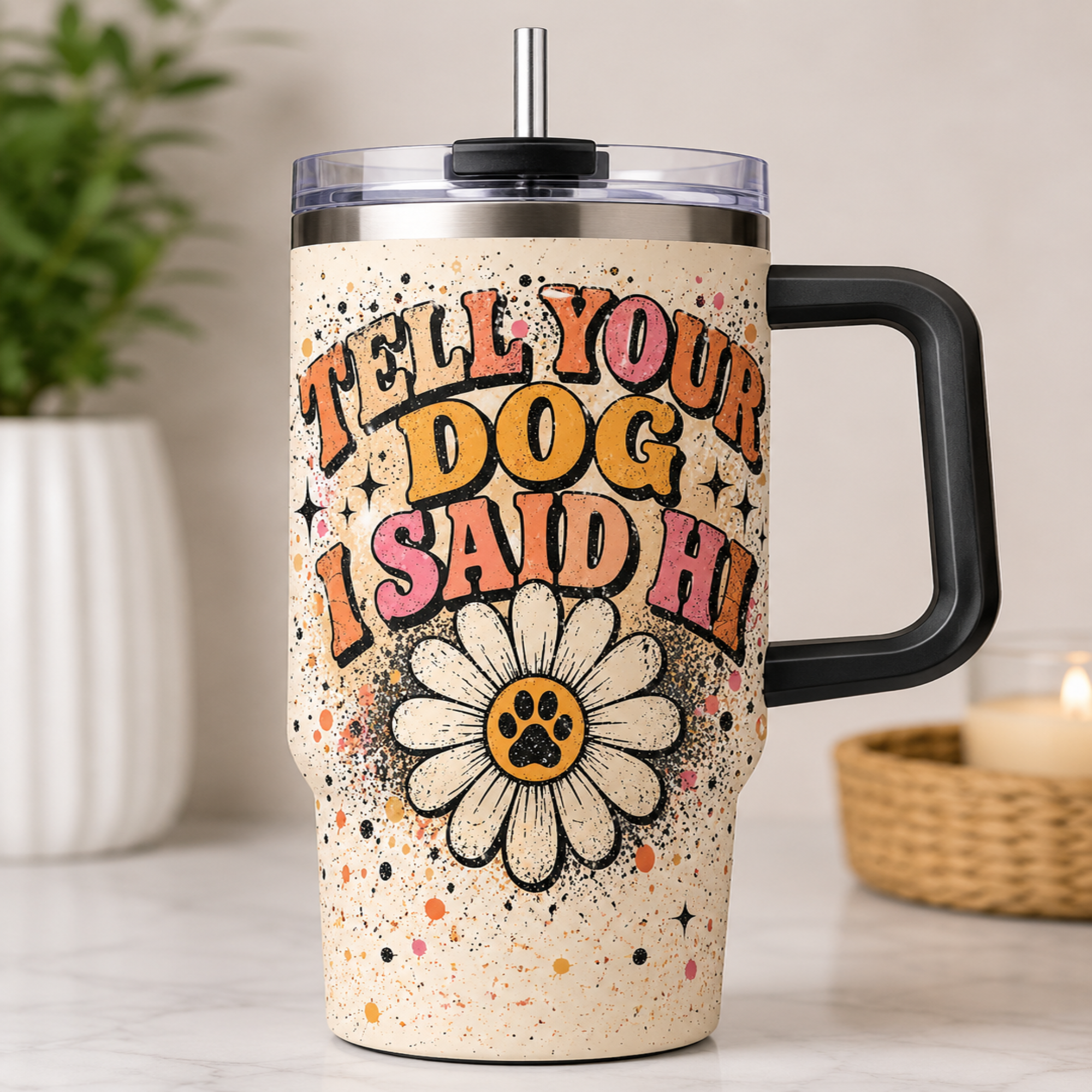 A stainless steel handled tumbler with a clear lid and straw, featuring a beige speckled background and the text 'TELL YOUR DOG I SAID HI' above a daisy flower with a paw print center. The tumbler is shown on a marble countertop with a blurred plant and candle in the background.