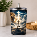 A clear-lidded stainless steel tumbler with a metal straw, featuring a printed religious scene with a lion, a lamb, a crucifix, clouds, and a glowing cross. The tumbler is photographed on a light countertop with a blurred plant and candle in the background.