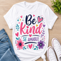 A white graphic t-shirt with the Spanish phrase 'Be Kind SÉ AMABLE' in pink and navy lettering, surrounded by colorful floral and heart illustrations. The t-shirt is styled in a flat-lay arrangement on a light wood background, accompanied by blue jeans, white sneakers, and a small green plant.