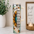 A handled stainless steel tumbler with a printed inspirational Bible verse design and a large sunflower graphic, styled on a marble countertop with a potted plant and a framed sign in the background.