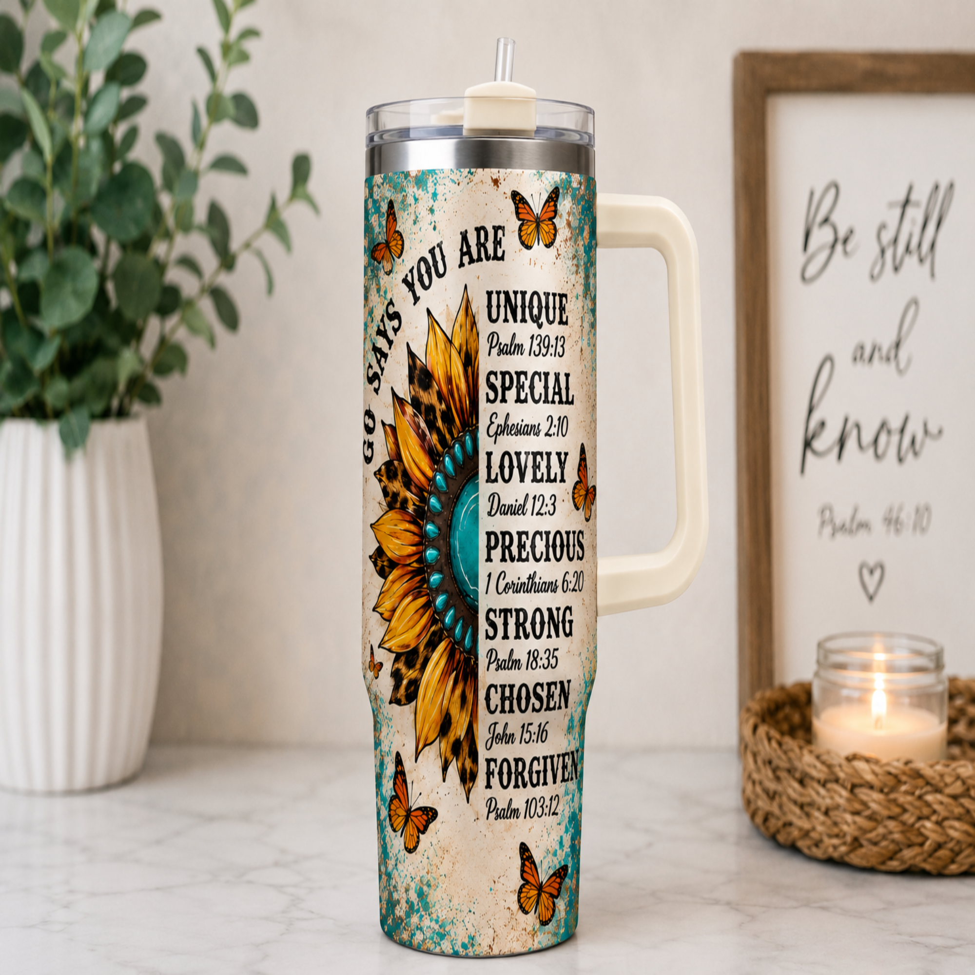 A handled stainless steel tumbler with a printed inspirational Bible verse design and a large sunflower graphic, styled on a marble countertop with a potted plant and a framed sign in the background.