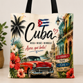 A canvas tote bag featuring a colorful Cuban-themed illustration with the word 'Cuba' in large script, a red classic car, tropical flowers, palm trees, and colonial-style buildings. The bag has black handles and is photographed against a light neutral background with a small plant partially visible on the left.